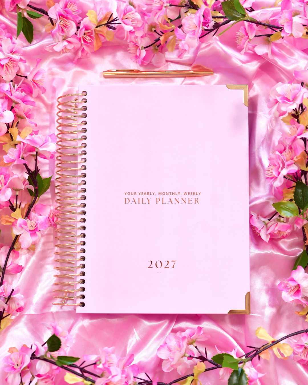 Pink 2027 Daily Spiral Planner with gold spiral binding among pink blossoms