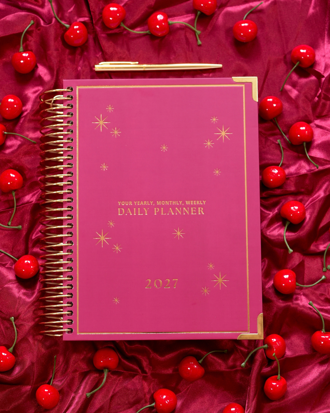 Burgundy 2027 Daily Spiral Planner with gold star details on a red satin background with cherries
