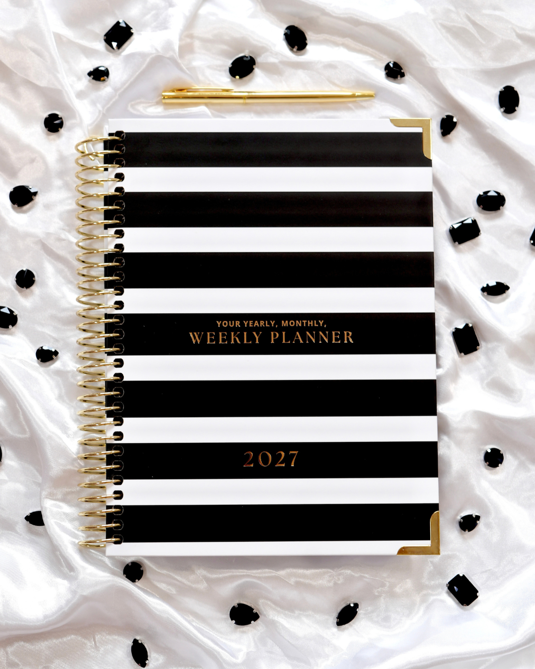 Black and white striped 2027 Weekly Spiral Planner with gold spiral binding and a gold pen on white satin