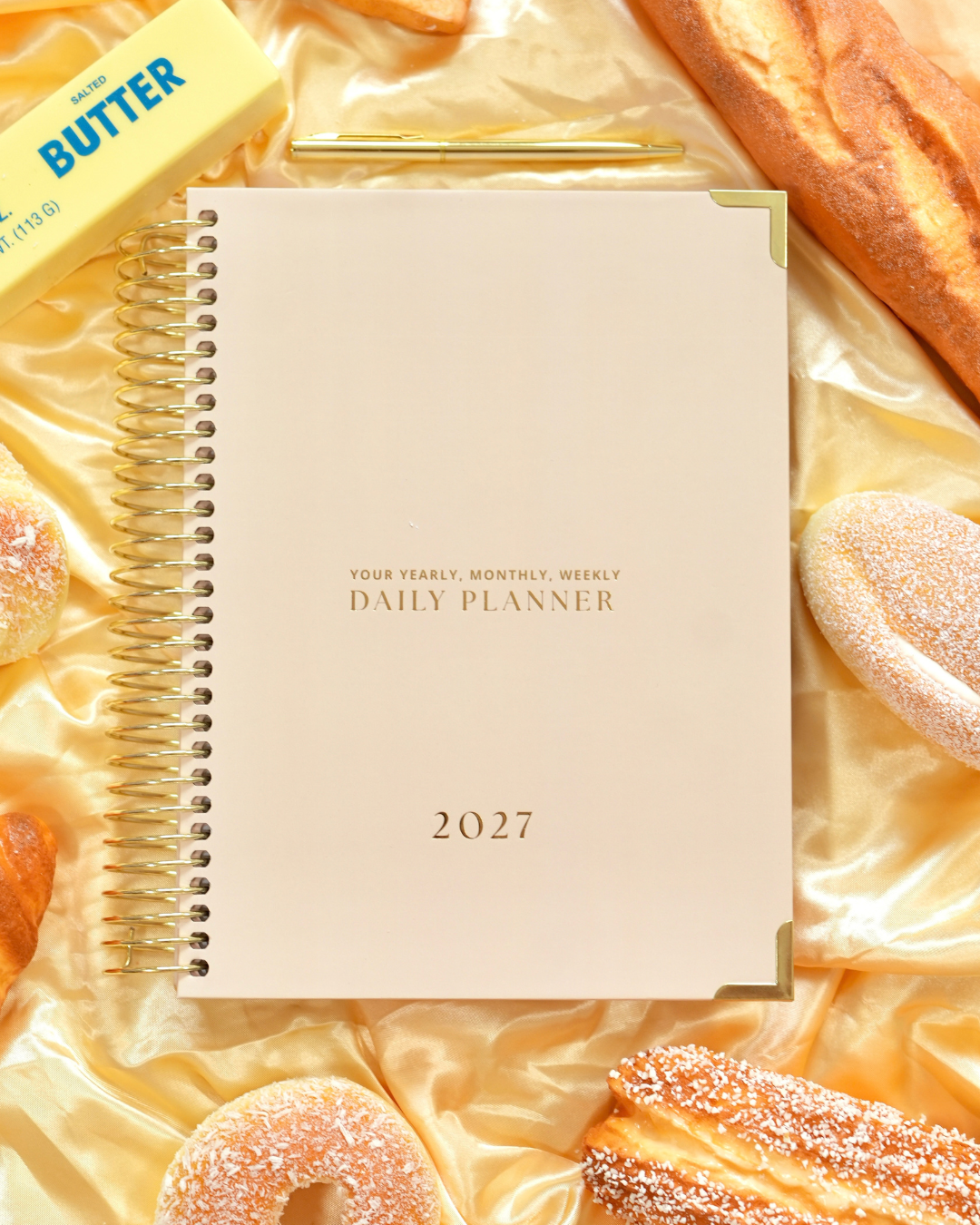 Beige 2027 Daily Spiral Planner with gold spiral binding beside butter, bread, and pastries