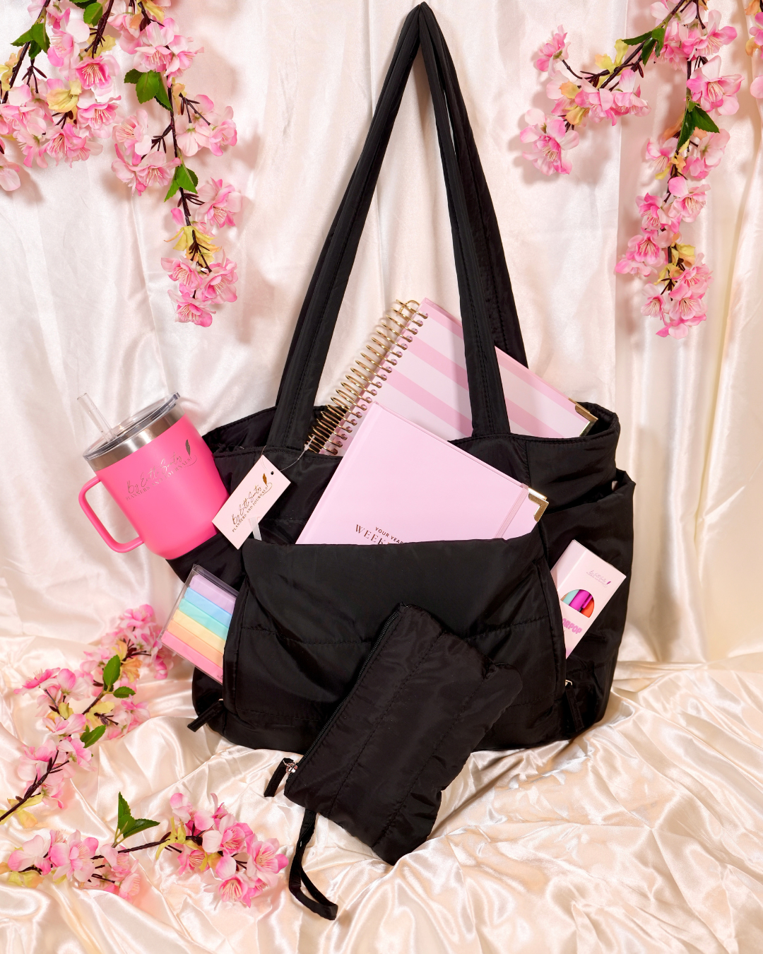 Black Travel Tote holding a planner, a pink tumbler, and a pouch on cream satin