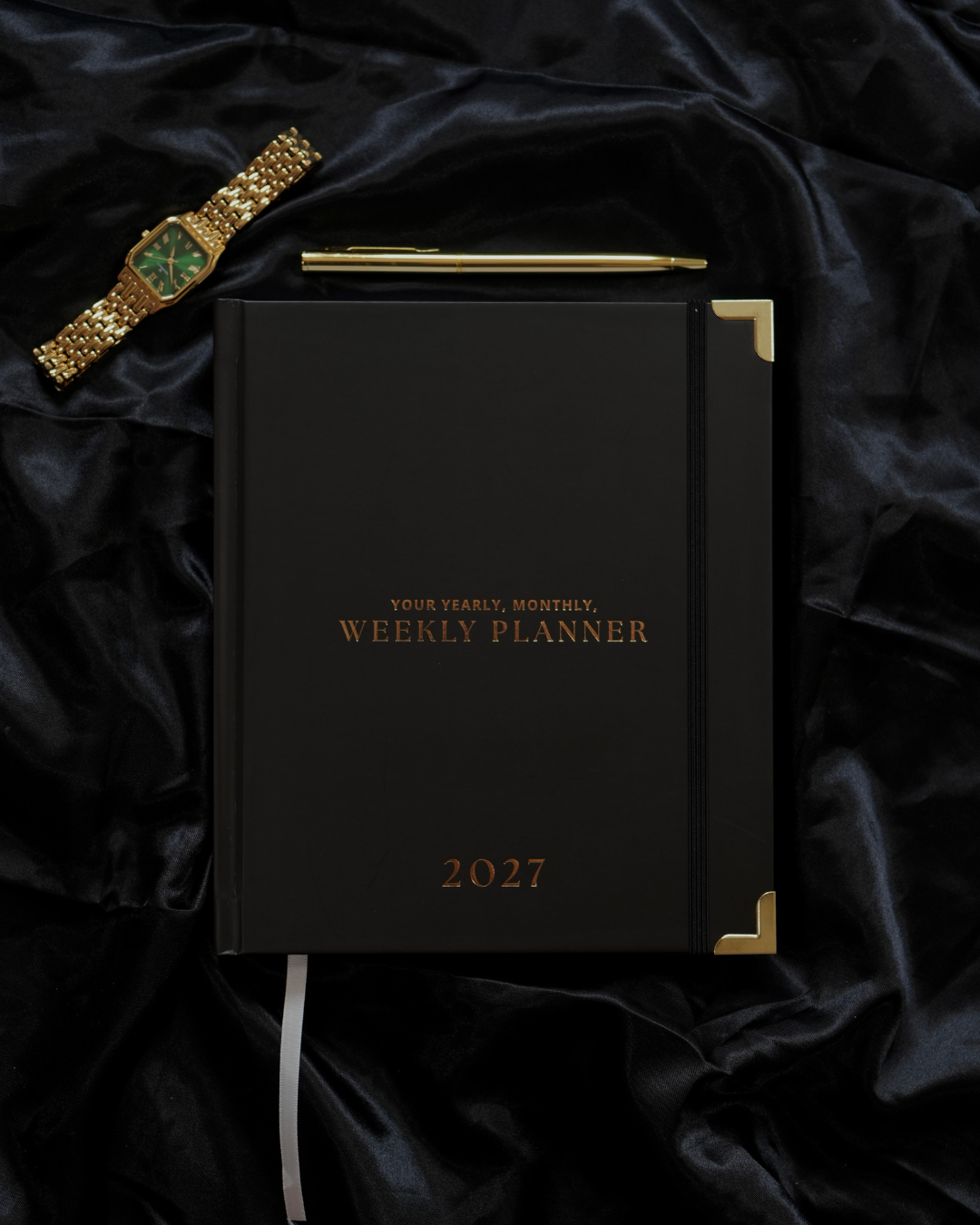 Black 2027 Weekly Mini Planner with gold corners, a gold pen, and a gold watch on black velvet